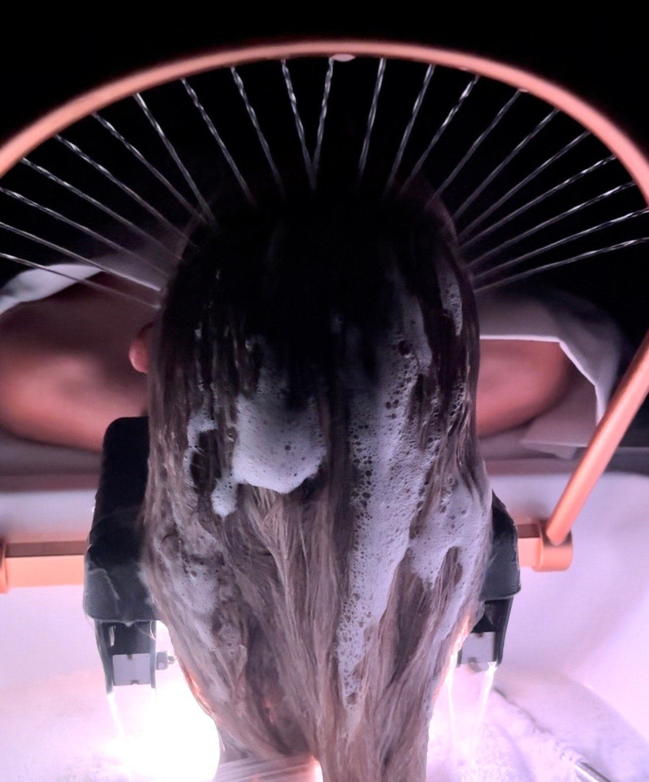Back view of a woman with long brown hair under a halo waterfall during a Japanese Head Spa treatment. Back view of a woman with long brown hair under a halo waterfall during a Japanese Head Spa treatment.
